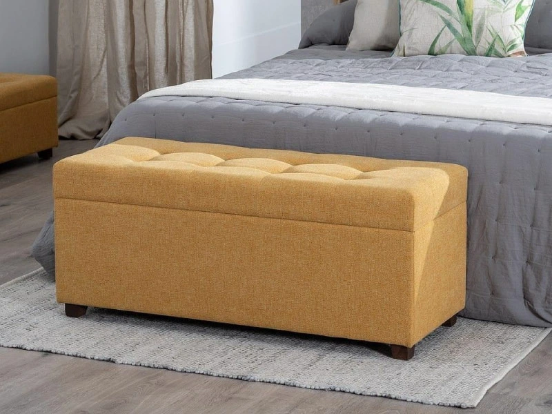 Baúl banqueta tapizado en tela mostaza al pie de una cama