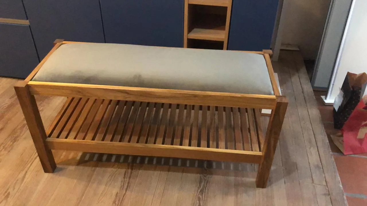 Banco de madera con asiento tapizado en tela verde grisácea y estante inferior